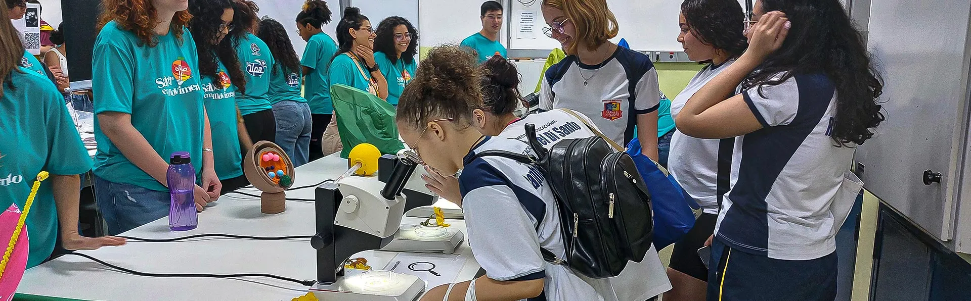 Grupo de pessoas em uma sala de aula usando camisetas turquesa com logotipo, reunidas em torno de uma mesa branca com equipamentos científicos incluindo microscópio, objetos amarelos e recipientes brancos, enquanto uma pessoa em primeiro plano manipula um equipamento com câmera fotográfica pendurada no pescoço, com observadores ao fundo próximos a um quadro branco e janela com vista para área externa.